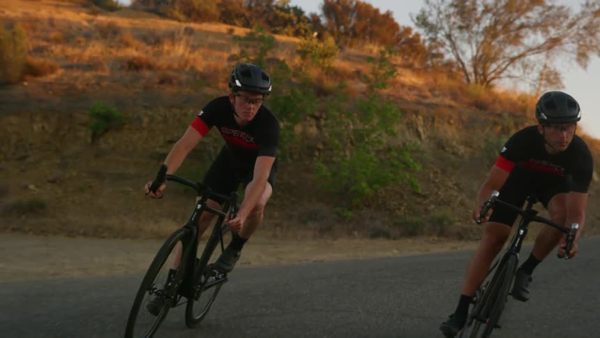 Two men ride bicycles on the road near a hill under a blue sky