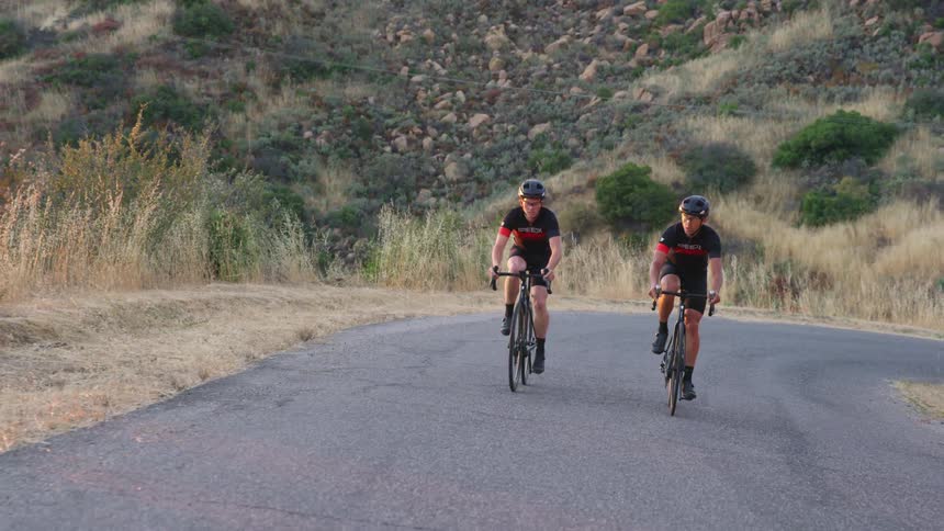 Two bicyclists ride down a road past a field and hills in a rural area
