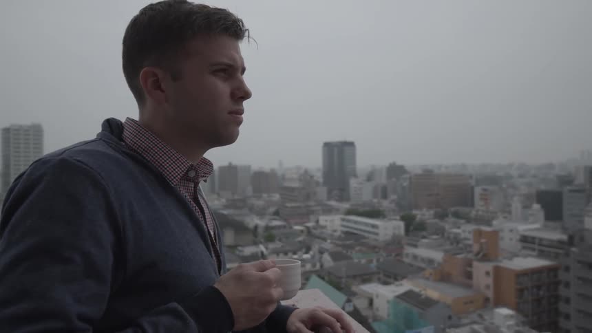 A man is watching a city from a balcony as he holds a mug
