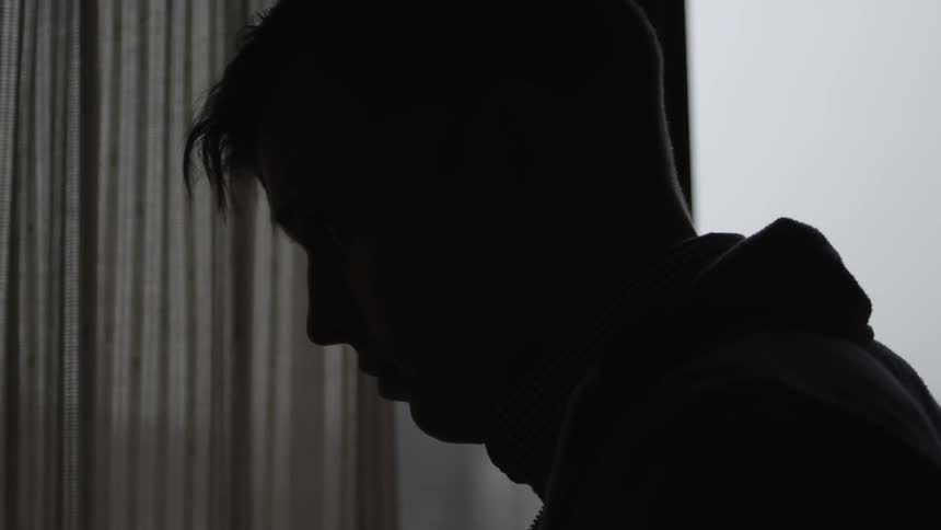 A man is looking down in a dark room by a window