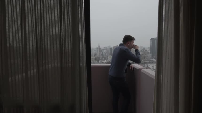 A man stands on a balcony sipping from a coffee cup