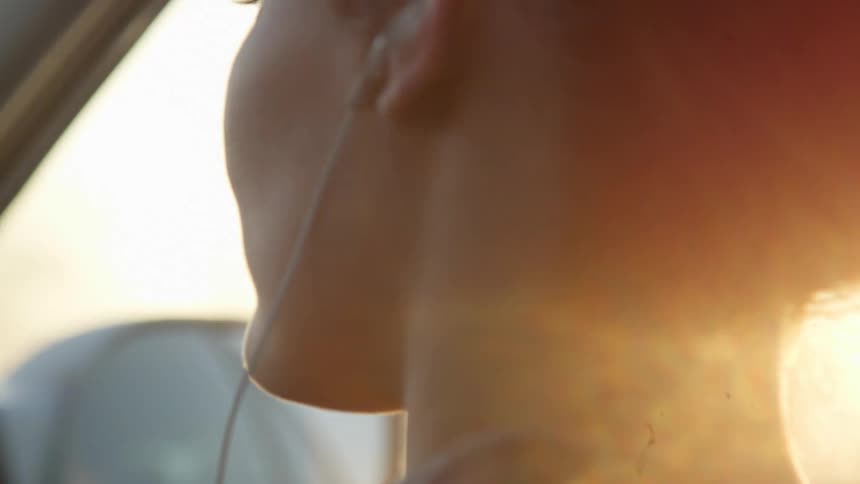 Sunlight flares around the head of a white woman sitting in a moving car and wearing ear buds