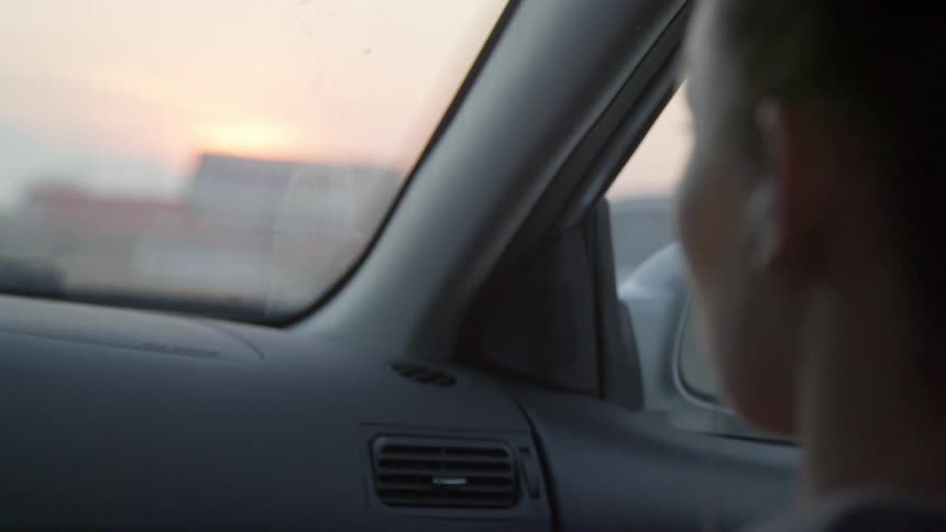 Woman with white earphones sits in a car driving across the road at sunset