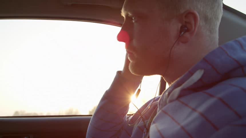 A white man sits in the back seat of a car and puts an ear bud into this ear