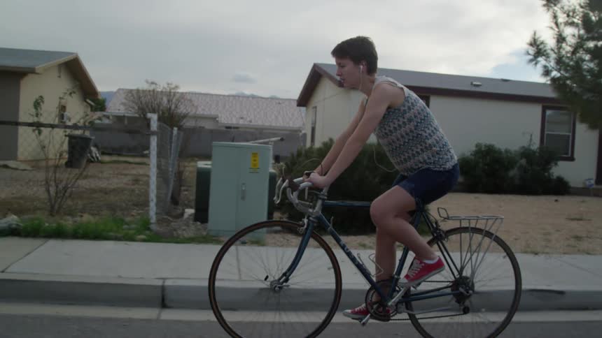 Female wearing earbuds while riding a 10 speed bicycle in the road next to a sidewalk through a neighborhood