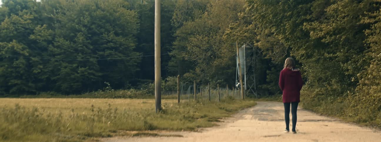 Woman with blonde hair and red sweater walks along a countryside road next to trees