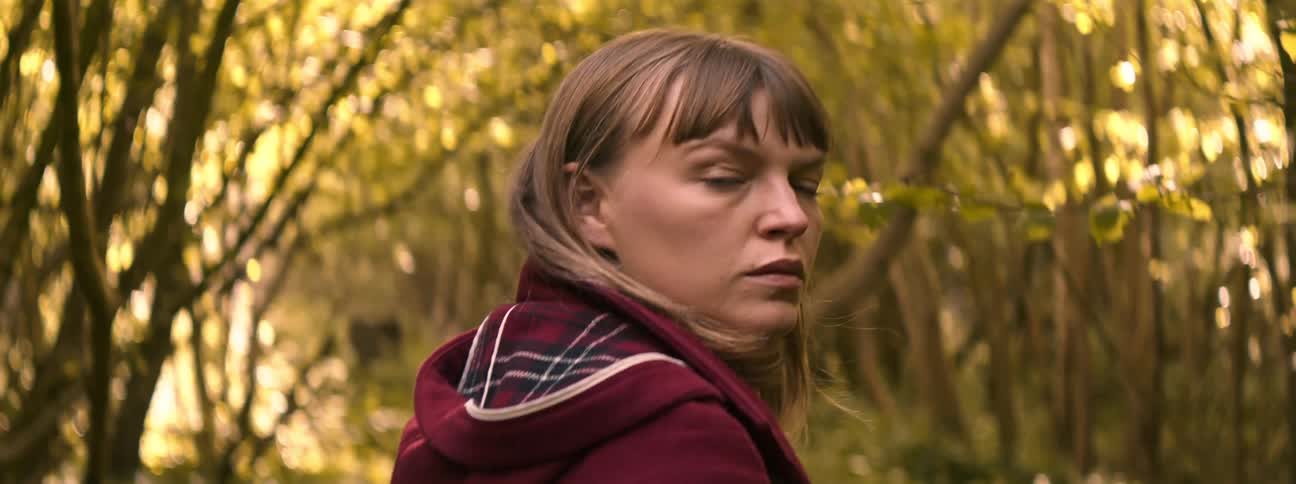 A white woman in a coat with a hood turns her head around in a forest