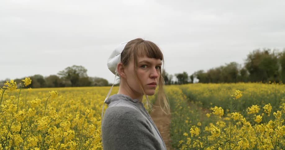 A young woman wearing a bonnet stands in a field among rows of flowering plants and then turns to leer at the camera