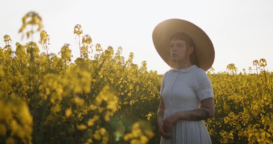 A woman stands alone wearing a dress and sun hat in a field of yellow flowering plants