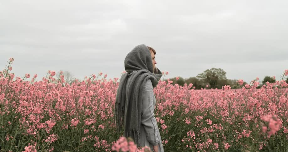 A woman wearing a dress and a scarf wrapped around her head stands in a field of pink flowers