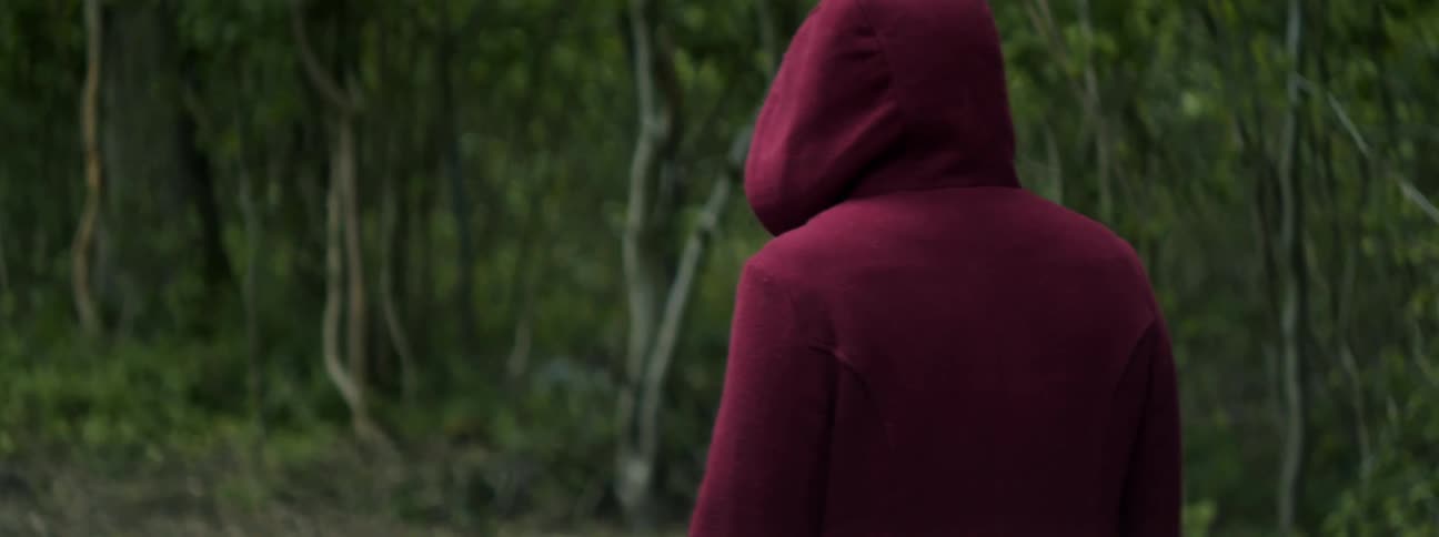A person with their face hidden by a maroon hood walks through a woodland during the day