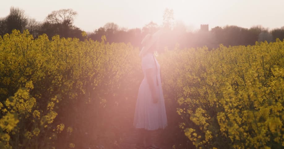 Woman wearing white dress and a sun hat stand on a field of yellow wild flowers