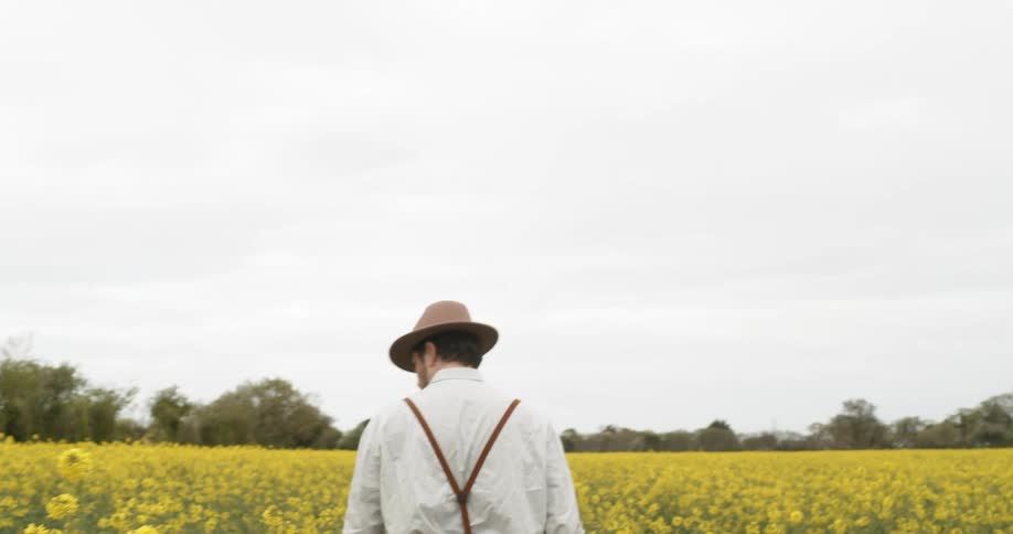 A man wearing a white shirt and suspenders walks in a field of yellow mustard flowers