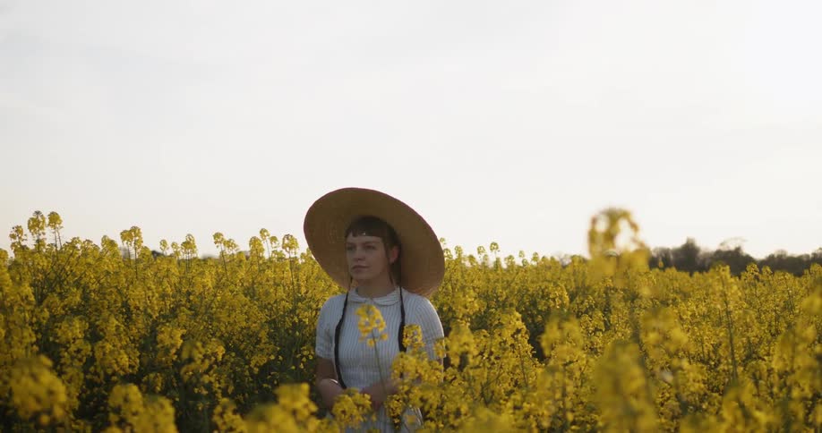 A woman wearing a white dress and a big brim hat walks through a field of flowering mustard plants