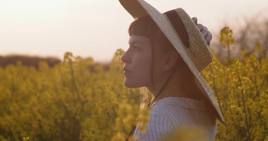 A woman wearing a sun hat stands alone in field of yellow flowering plants at dusk