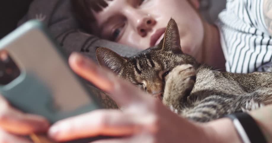A white female child lies in bed with a cat while looking at her smartphone