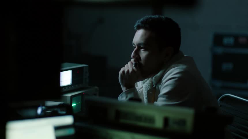 A man sitting at a desk filled with machines leans forward and puts his hand to his mouth as he watches a monitor