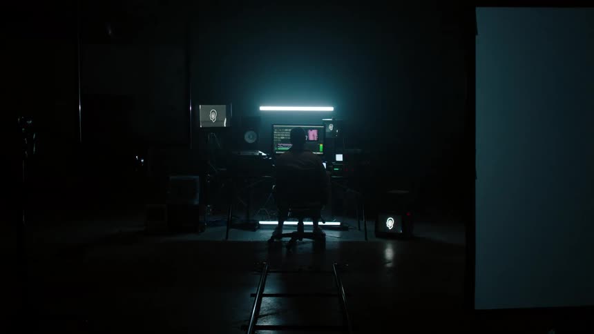A man sits in a chair at a desk and looks at a computer monitor in a room filled with tech devices