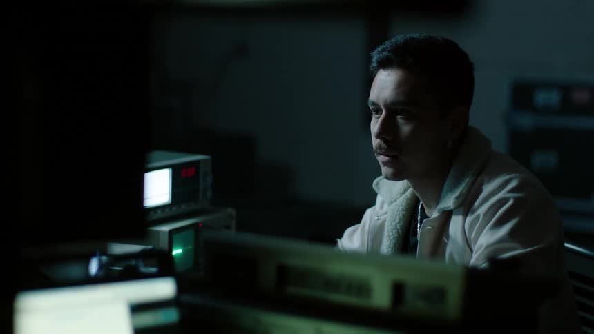 A man sits in a dark room looking anxiously at a set of old electrical equipment before clasping his hands to pray