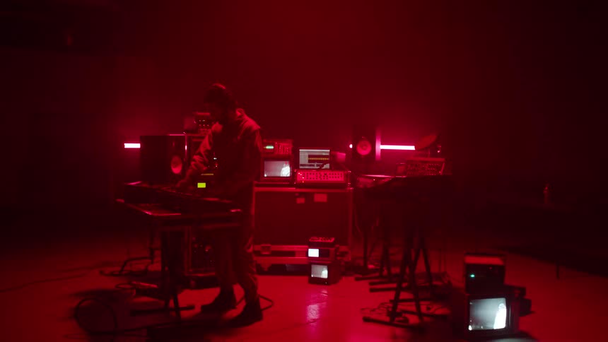 Musician play keyboards under flashing red lights surrounded by electronic equipment