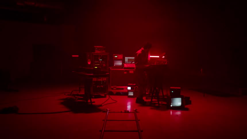 A person adjusting a device while standing surrounding by monitors showing static in a dark room illuminated by a rapidly flashing red light