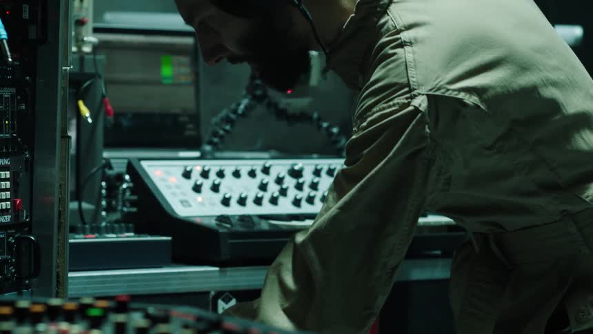 A man with headphones touching a musical keyboard surrounded by equipment and smoke in the background