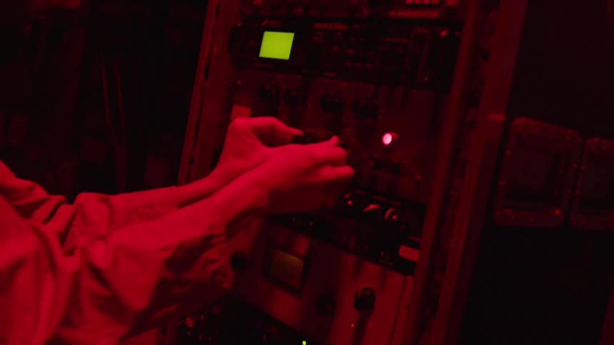 A man wearing headphones adjusts knobs and dials as red lights flash and glow