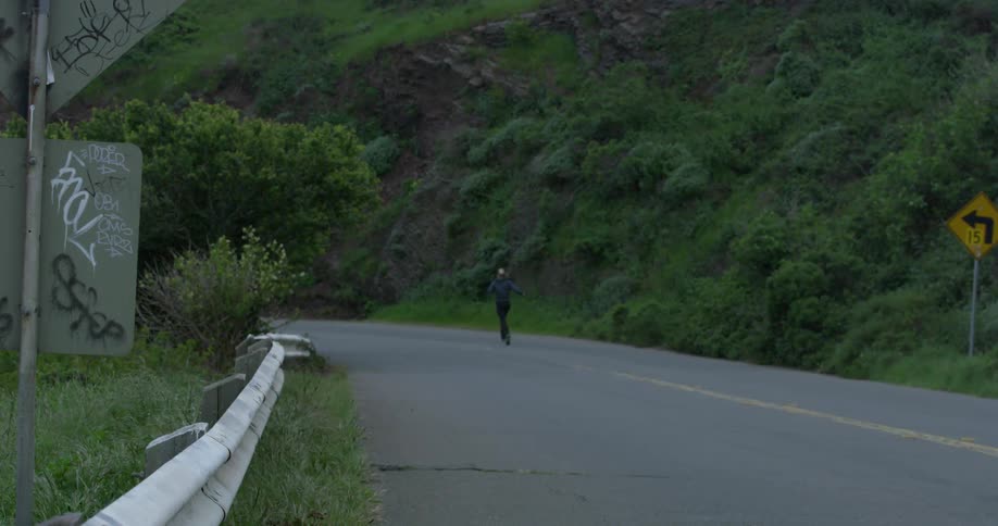 A blonde woman jogs down a road next to a steep hill