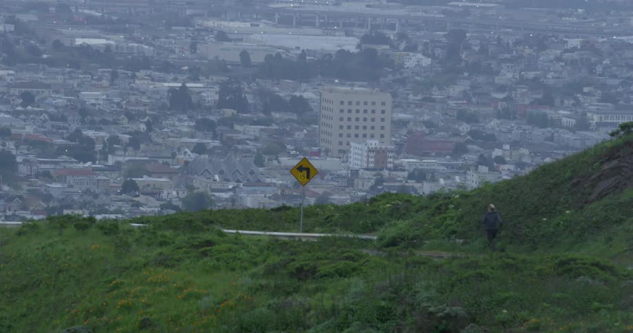 A jogger runs down a road next to a hill within site of a large downtown part of a city