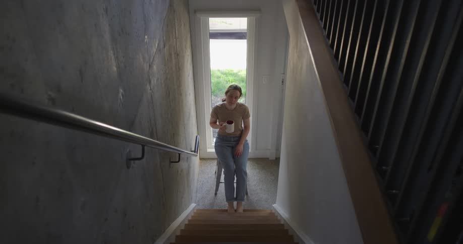 A woman sits on a chair at the bottom of a staircase drinking from a mug