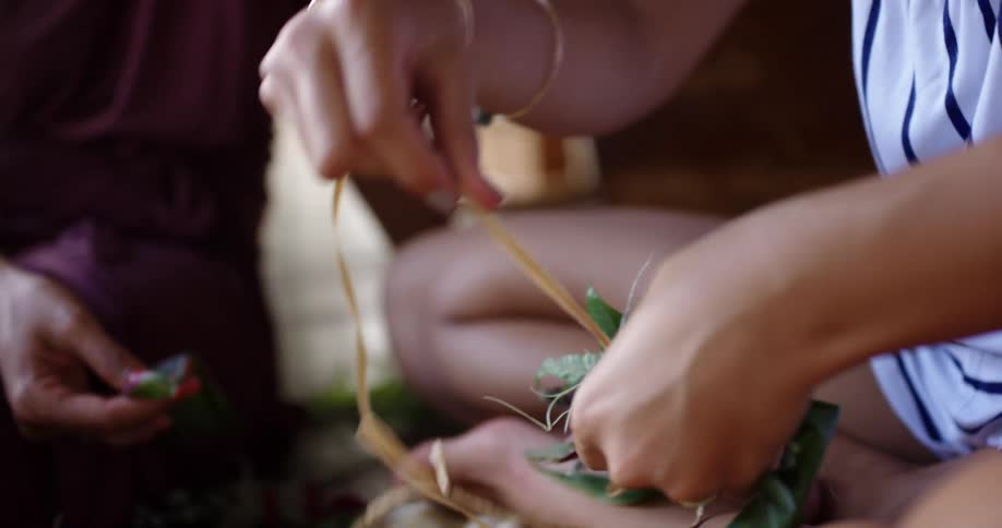 Two women sit on a floor as one of them wraps string around a small bouquet of plants and flowers