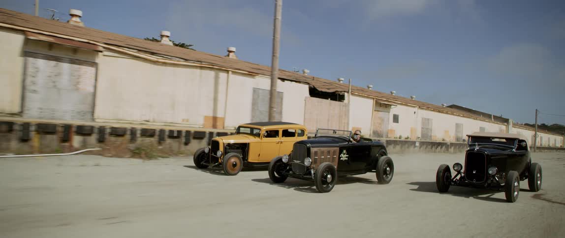 A trio of vintage vehicles traveling down a wide paved area next to a long dilapidated building