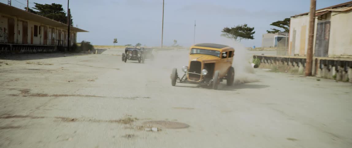 Vintage cars race on a dusty dirt road next to some buildings