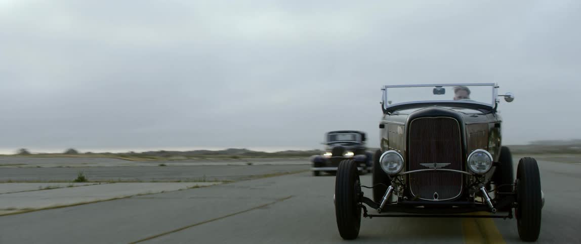 Three vintage cars drive along road through empty fields under overcast sky