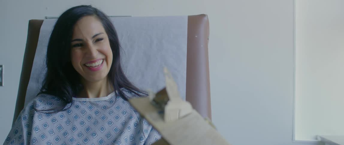A woman in a hospital gown sitting on a chair and smiling while the doctor writes on a clipboard