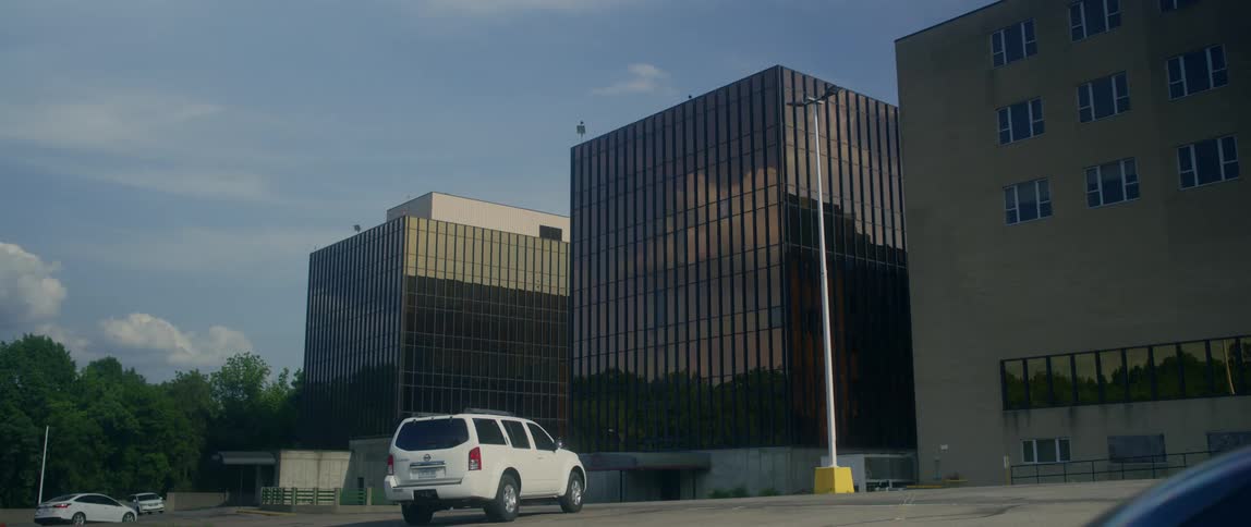 Sky clouds and trees are reflected on the glass facade of an office building near a parking lot with only a few cars