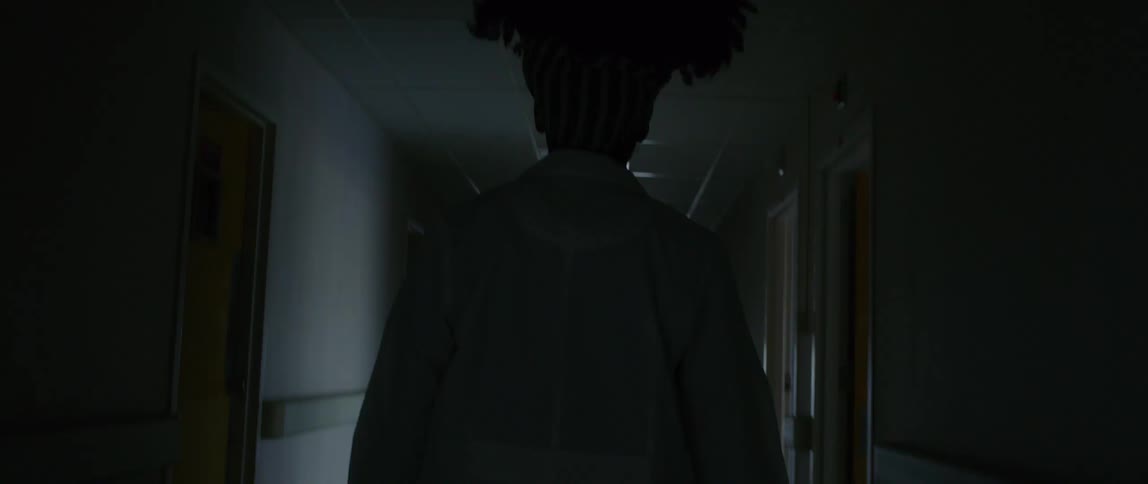 A hospital staff member in scrubs walks down a dark hallway towards a bright doorway