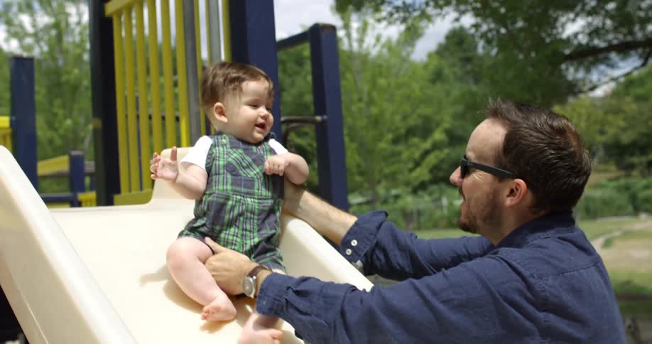 A man holds and slides a toddler down a slide in a backyard play area
