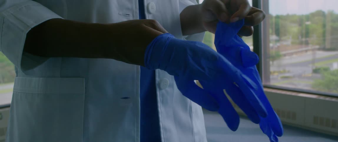 Medical practitioner dons gloves standing near window showing trees and buildings