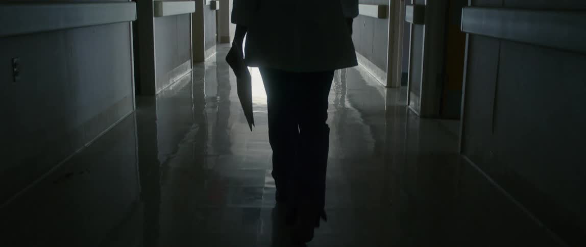 A female doctor walks down a dark corridor in a hospital while carrying a clipboard