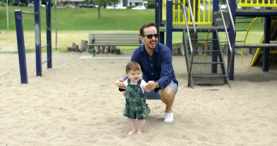 A father holds the hands of a baby standing in the sand at a public playground
