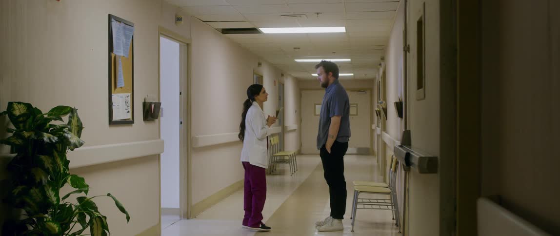 A doctor walks down a hallway and a man stands up to listen to her