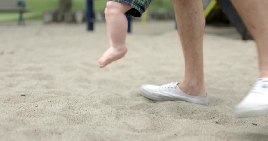 A man helping a smiling baby to hop through the sand of a playground by repeatedly lifting the baby when its feet touch the ground