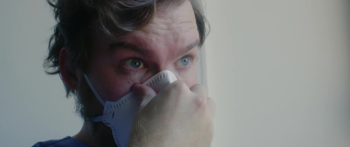 A white male wearing scrubs carefully puts on a face mask and walks away