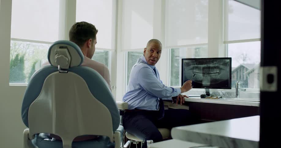 Dentist sitting in front of patient in dental chair discusses the patients x ray on the monitor and points to it using a pen as he speaks
