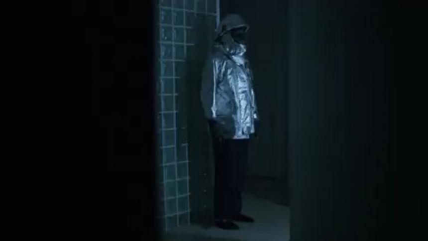 A figure clothed in silver clothing and a mask stands in a hallway as lights flash