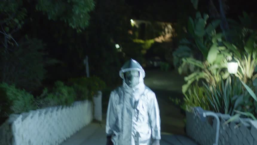 A man stands on a walkway with fists clenched and wearing a mask and silver coat as lights flash around him