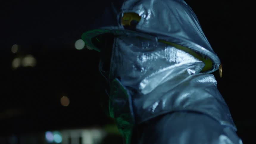 A man dressed in some type of protective suit and face mask stands outdoors at night with lights flashing