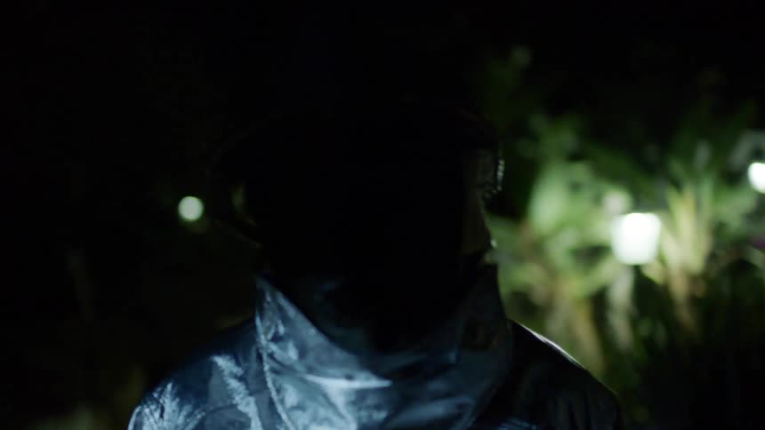 Person wearing full face mask stands revealed by flashing lights near house yard