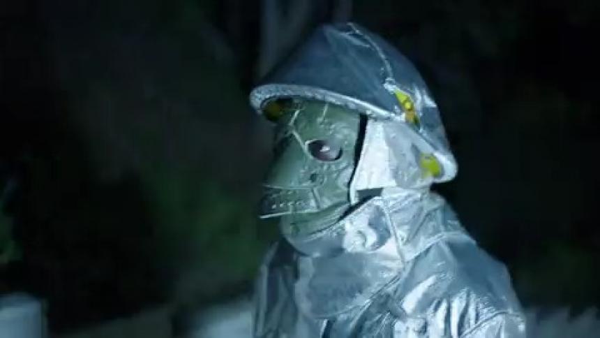 A person covered in a bizarre mask and reflective clothing stands outdoors at night